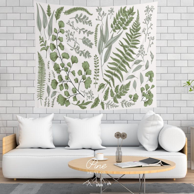 May include: A white tapestry with a green botanical print featuring various types of leaves and ferns. The tapestry is hanging on a white tiled wall above a white couch with white pillows. The text "Pine Print Co." is printed in the bottom center of the tapestry.