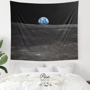 May include: A rectangular wall tapestry featuring a high-resolution space photograph of the gray, cratered lunar surface in the foreground with the blue and white Earth rising above the horizon against a deep black sky. The fabric is draped.