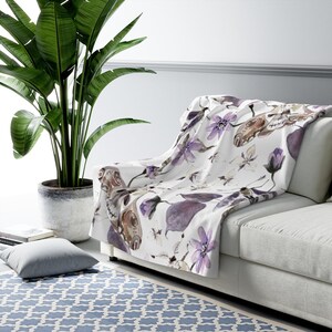 May include: A white throw blanket with a watercolor pattern of purple flowers and butterflies. The blanket is draped over a white couch.