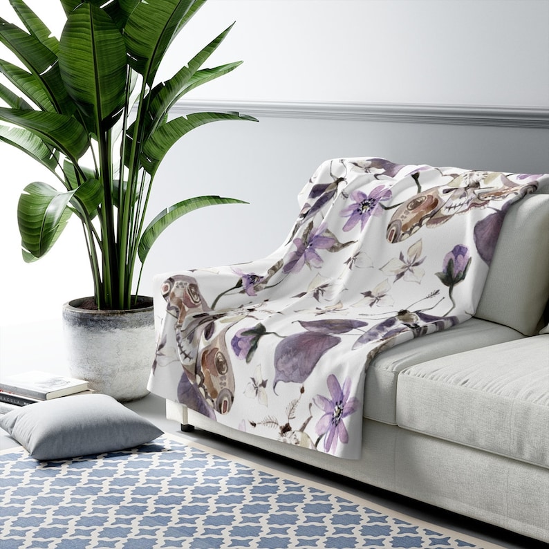 May include: A white throw blanket with a watercolor pattern of purple flowers and butterflies. The blanket is draped over a white couch.