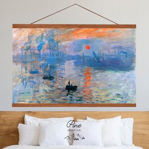 May include: A canvas print of Claude Monet's Impression, Sunrise, depicting a hazy sunrise over a harbor with ships and a small boat in the foreground. The print is framed with a wooden frame and hanging on a wall above a bed with white pillows.