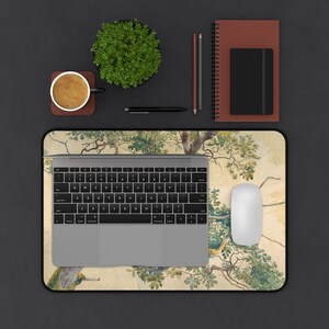 May include: A laptop computer with a gray keyboard and trackpad sits on a desk mat with a watercolor design of trees and birds. A coffee cup, a potted plant, pencils, and a notebook are also on the desk.