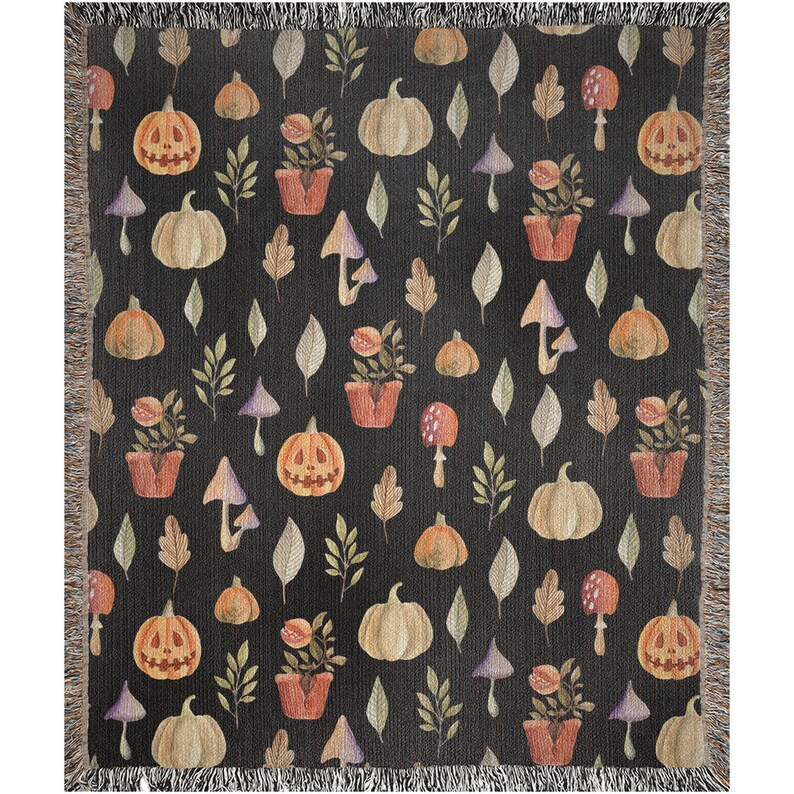 May include: A black woven throw blanket with a repeating pattern of orange pumpkins, green leaves, and purple mushrooms. The pumpkins have jack-o'-lantern faces.