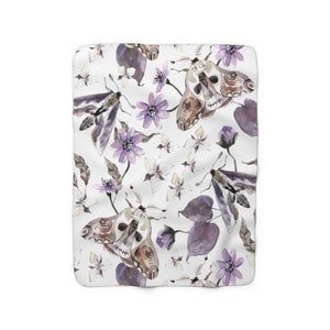 May include: A white fleece blanket with a repeating pattern of purple flowers, leaves, and moths with skull-like faces. The moths are brown and gray with purple accents.