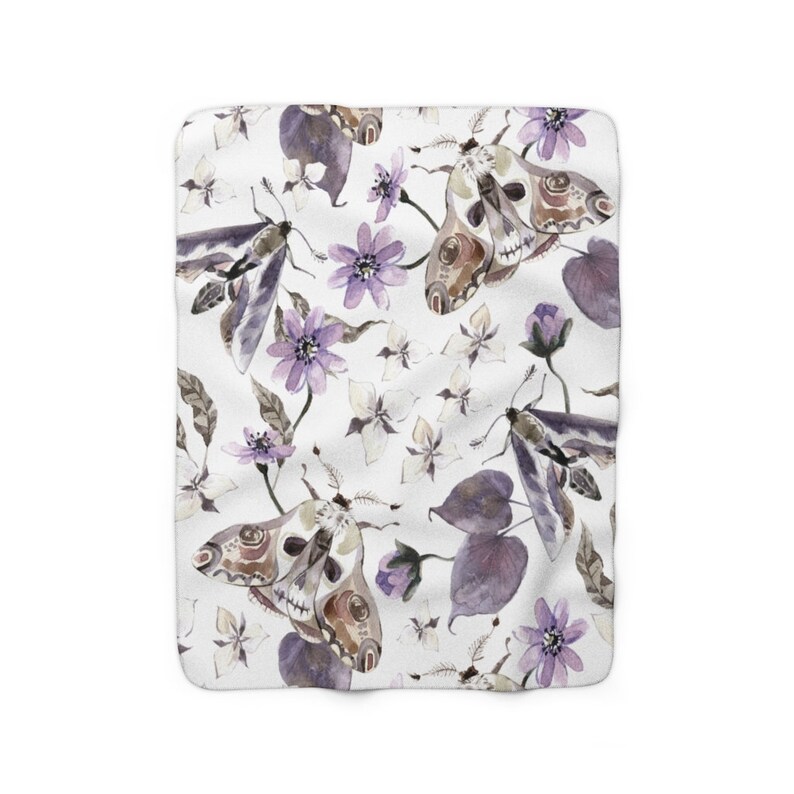 May include: A white fleece blanket with a repeating pattern of purple flowers, leaves, and moths with skull-like faces. The moths are brown and gray with purple accents.