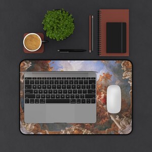 May include: A black and white laptop computer with a keyboard and trackpad is on a desk mat with a colorful painting of a sky with clouds and angels. A white mouse is on the desk mat. There is a potted plant, a cup of coffee, two pencils, and a pen on the desk.