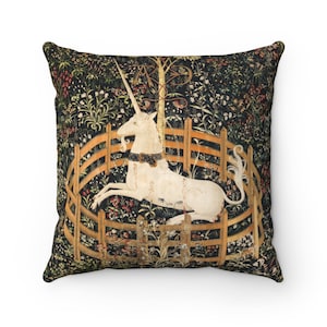 May include: A square throw pillow with a tapestry design featuring a white unicorn trapped in a circular fence. The unicorn is lying down with its head turned to the side. The background is a dark green with a variety of flowers and plants.
