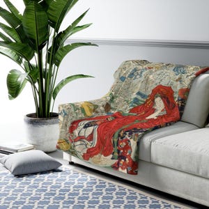 May include: A rectangular throw blanket featuring a detailed Japanese woodblock print illustration. The design depicts a central figure draped in a vibrant red robe, surrounded by skeletal figures and intricate patterns in muted tones of beige and grey.
