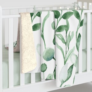 Puede incluir: Una cuna blanca con una manta de estampado floral verde y blanco drapeada sobre el lado. La manta est&aacute; hecha de tela suave y mullida y presenta un dise&ntilde;o de estilo acuarela.