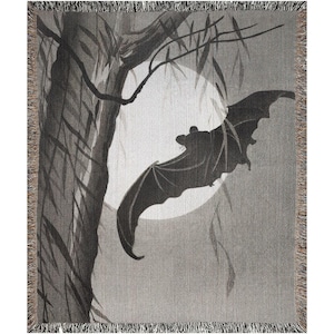 May include: A woven throw blanket featuring a grayscale illustration of a bat in flight against a large, circular full moon. The scene includes the silhouette of a tree trunk and hanging branches on the left side, finished with a fringed edge.