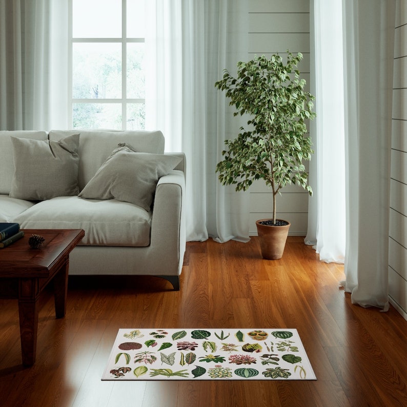 May include: A white area rug with a botanical print featuring various green leaves and flowers. The rug is placed on a hardwood floor in a living room setting with a white sofa and a potted plant.