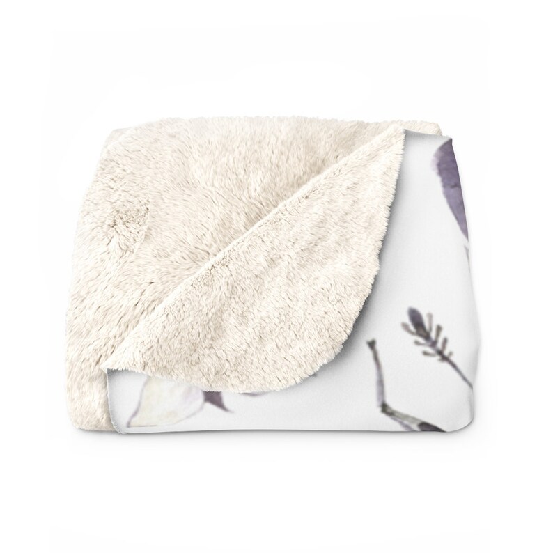 May include: A white faux fur throw blanket with a white and purple floral print. The blanket is folded in half and the faux fur side is facing the camera.
