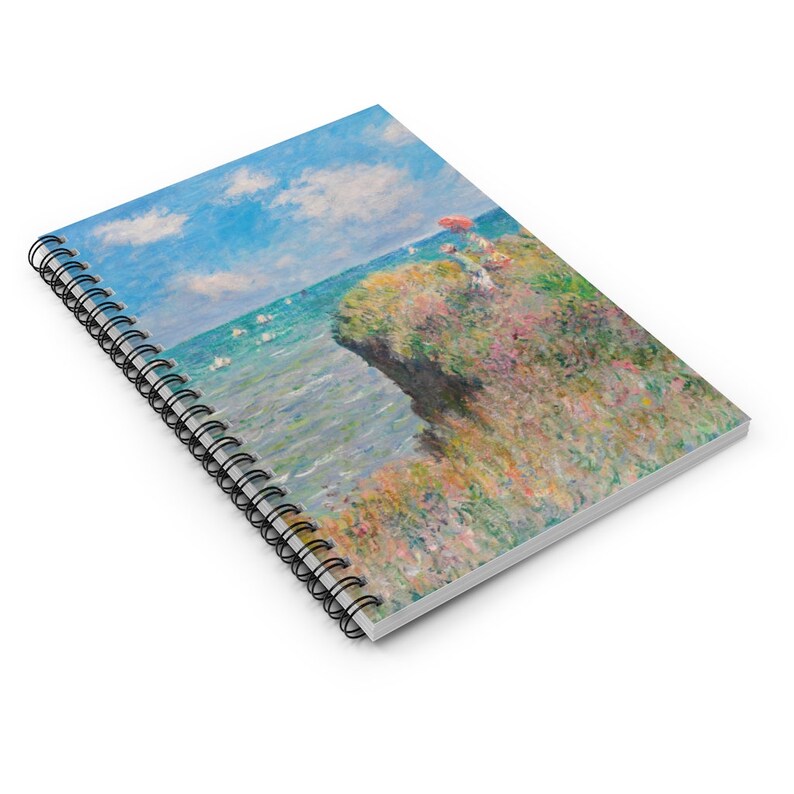 May include: A spiral bound notebook with a painting of a seascape. The painting features a rocky cliff with a small red umbrella and a view of the ocean with sailboats in the distance. The sky is blue with white clouds.