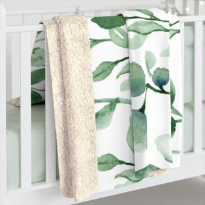 Puede incluir: Una manta de forro polar suave con estampado blanco y verde y un respaldo de sherpa crema. La manta est&aacute; doblada y drapeada sobre una cuna blanca.