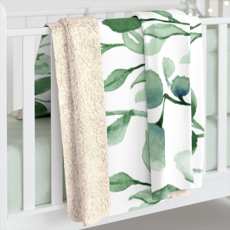 Puede incluir: Una manta de forro polar suave con estampado blanco y verde y un respaldo de sherpa crema. La manta est&aacute; doblada y drapeada sobre una cuna blanca.