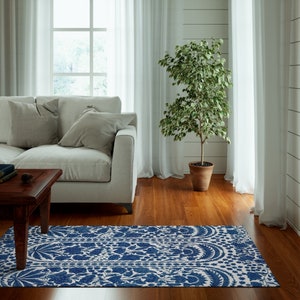 May include: A blue and white patterned rug with a floral design is placed on a hardwood floor. The rug is in front of a white couch and a dark wooden coffee table. A potted plant and white curtains are also in the room.