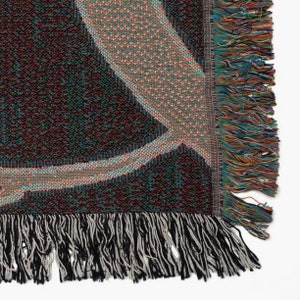 Puede incluir: Primer plano de una manta tejida con un dise&ntilde;o geom&eacute;trico en tonos negro, verde azulado y beige. La manta tiene un borde de flecos en hilos negros, grises y multicolores. La textura de la manta es visible.