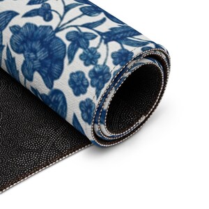 May include: Rolled-up rug featuring a blue floral pattern on a white background. The design includes stylized flowers and leaves. The rug's edges are neatly finished with a contrasting border, and the underside is black.