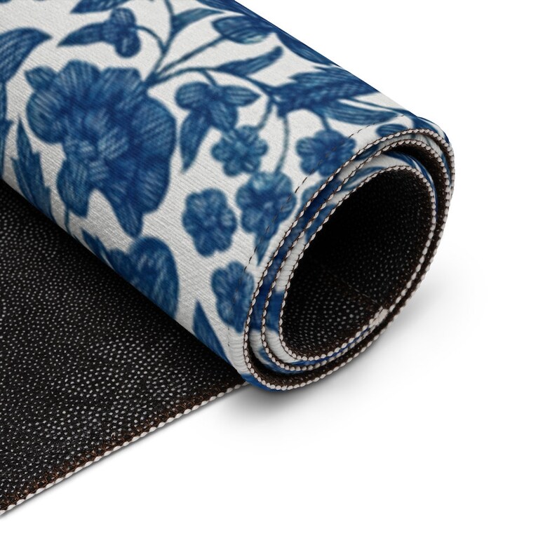 May include: Rolled-up rug featuring a blue floral pattern on a white background. The design includes stylized flowers and leaves. The rug's edges are neatly finished with a contrasting border, and the underside is black.