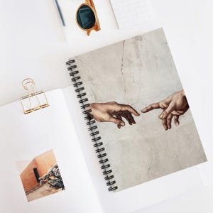 以下が含まれることがあります： 表紙にアダムの創造の複製が描かれたスパイラル綴じのノート。側面には金色の金属製バインダークリップが留められており、開いた白いページには砂漠の建物の小さな正方形の写真が置かれています。