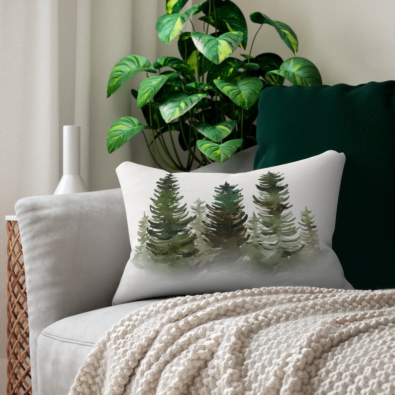 Puede incluir: Coj&iacute;n rectangular decorativo con una escena de bosque en acuarela con &aacute;rboles verdes y marrones sobre un fondo blanco. El coj&iacute;n est&aacute; colocado sobre un sof&aacute; gris con una manta de punto color crema.