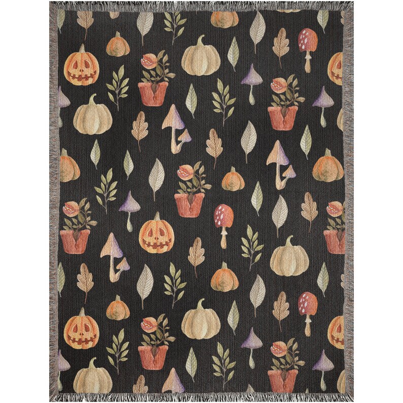 May include: A black woven throw blanket with a repeating pattern of pumpkins, mushrooms, leaves, and potted plants. The pattern is in shades of orange, brown, green, and white.