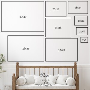 May include: A white wall with a variety of black framed prints of different sizes. The prints are labeled with their dimensions in inches. The wall is above a wooden bench with white pillows.