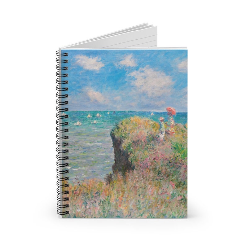 May include: A spiral bound notebook with a painting of a seascape. The painting depicts a rocky cliff with a couple sitting on it, looking out at the ocean. There are sailboats in the distance and a bright blue sky with white clouds.