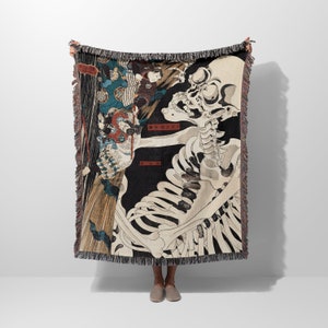 May include: A woven tapestry throw blanket featuring a black and white skeletal figure with a Japanese design. The skeleton is holding a sword and is surrounded by other figures in the background. The text "皆 皆 皆" is written in red on the blanket.