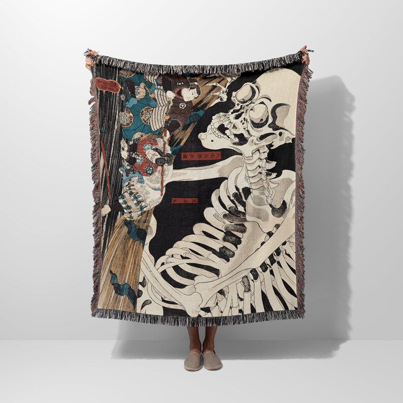 May include: A woven tapestry throw blanket featuring a black and white skeletal figure with a Japanese design. The skeleton is holding a sword and is surrounded by other figures in the background. The text "皆 皆 皆" is written in red on the blanket.