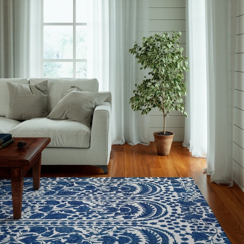 May include: A living room scene featuring a white sofa with gray pillows, a dark wood coffee table, and a patterned blue and white rug. A potted plant and sheer white curtains frame the window.