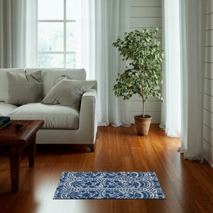 May include: A blue and white patterned rug is placed on a hardwood floor in front of a sofa. The rug has a detailed floral design. A potted plant and a window with white curtains are in the background.