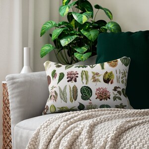 Puede incluir: Una almohada blanca con un estampado botánico que presenta varias hojas verdes y marrones. La almohada está en un sofá gris claro con una manta blanca.