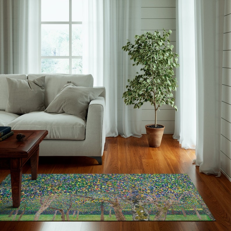 May include: A green, blue, and yellow area rug with a stylized painting of trees. The rug is in a living room with a white couch, a wooden coffee table, and a potted plant.