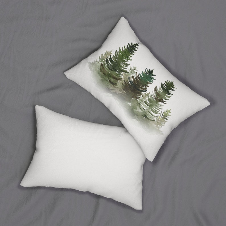 Puede incluir: Dos cojines rectangulares. Uno presenta una pintura de acuarela de pinos verdes sobre un fondo blanco. El otro coj&iacute;n es blanco liso. Los cojines est&aacute;n sobre una superficie gris.