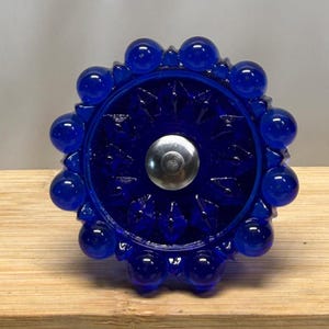 May include: A circular cobalt blue glass cabinet knob featuring a faceted starburst pattern in the center and a border of smooth, rounded glass spheres. A polished silver-toned metal screw head is centered in the middle of the blue glass knob.