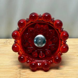 May include: A circular red glass knob featuring a faceted starburst pattern in the center and a ring of smooth, rounded spheres along the outer edge. A polished silver-toned metal bolt is centered in the middle of the red glass component.