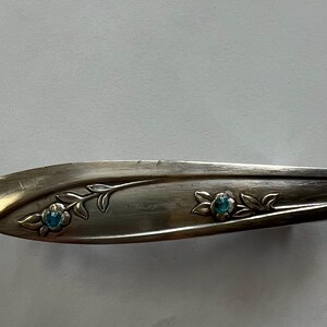 May include: Silver-coloured metal hair clip with floral and leaf design. The clip features two blue gemstone accents in the shape of flowers. The clip has a curved shape and is likely vintage.