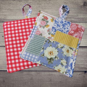 May include: Two handmade pot holders. One is red and white checkered, the other is a patchwork of floral and patterned fabrics in shades of blue, pink, yellow, and green. Each has a fabric loop for hanging.