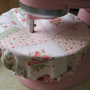 May include: A pink stand mixer with a floral patterned cover. The cover is made of patchwork fabric with pink roses, green leaves, and white backgrounds. The mixer is a light pink color.