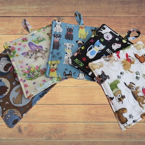 May include: A collection of four square fabric pot holders featuring various animal prints. One shows kittens and flowers, another blue with dogs, one black with cats and the word MEOW, and the last white with dogs and boots. Each has a fabric loop.