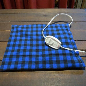 May include: A blue and black plaid heating pad with a white control and cord. The pad is rectangular and has a button on the side. The control has settings for different heat levels.