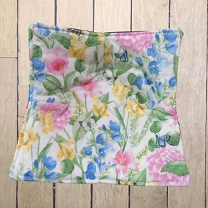 May include: A white quilted hot pad with a floral print featuring pink, yellow, and blue flowers and butterflies. The hot pad has a dark blue border.