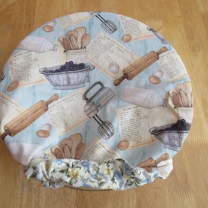 May include: A round fabric bowl cover featuring a light blue background with a repeating pattern of vintage kitchen tools, including rolling pins, whisks, colanders, and recipe cards. The edge has a gathered, floral-patterned elastic trim.