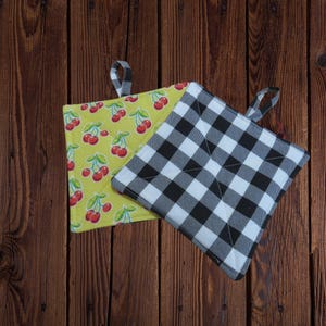 May include: Two square pot holders. One is yellow with a cherry pattern, and the other is black and white checkered. Both have a small loop for hanging. The pot holders are on a wooden surface.