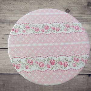 May include: A circular fabric cover featuring a repeating pattern of pink roses, white lace-like borders, and small white floral motifs on a soft pink background. The design is arranged in horizontal bands across the round textile surface.