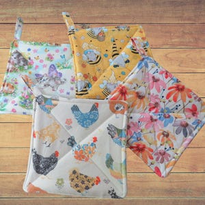 May include: A collection of five square pot holders with hanging loops. The pot holders feature various patterns, including kittens and butterflies, bees and gnomes, colorful flowers, and chickens. Each pot holder is made of fabric and has a quilted appearance.