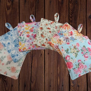May include: A collection of five square fabric pot holders featuring various floral patterns in shades of blue, pink, yellow, and green. Each quilted square includes a small fabric loop at the top corner for hanging, displaying distinct botanical prints.