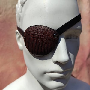 May include: A brown leather eye patch with a textured, crocodile-like pattern. The eye patch is secured with a black strap and a metal rivet. The eye patch is designed to cover one eye.
