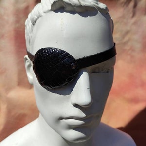 May include: A black leather eye patch with a crocodile-embossed design. The patch is worn over the right eye and secured with a strap.